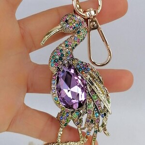 May include: A gold-toned bird keychain adorned with colorful rhinestones. The bird features a large, purple, teardrop-shaped crystal at its center. The keychain has a lobster clasp and a key ring.