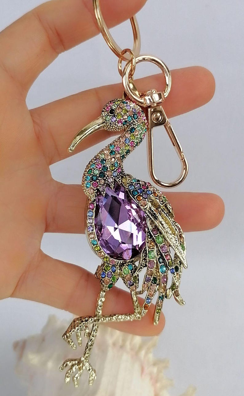 May include: A gold-toned bird keychain adorned with colorful rhinestones. The bird features a large, purple, teardrop-shaped crystal at its center. The keychain has a lobster clasp and a key ring.