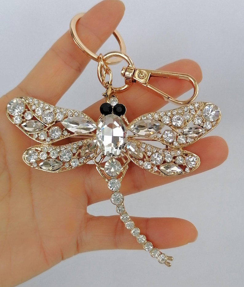 Op de afbeelding: Een goudkleurige sleutelhanger in de vorm van een libel met heldere kristallen accenten. De libel heeft een groot, centraal kristallen lichaam en vleugels versierd met kleinere kristallen. De sleutelhanger bevat een gouden ring en een sluiting.