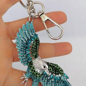 May include: A silver-toned eagle keychain with teal and green jewel-like accents. The eagle's wings are spread, and the keychain has a clasp and a ring for keys. The eagle's head is silver, and the body is adorned with sparkling gems.
