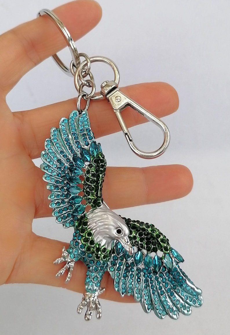 May include: A silver-toned eagle keychain with teal and green jewel-like accents. The eagle's wings are spread, and the keychain has a clasp and a ring for keys. The eagle's head is silver, and the body is adorned with sparkling gems.