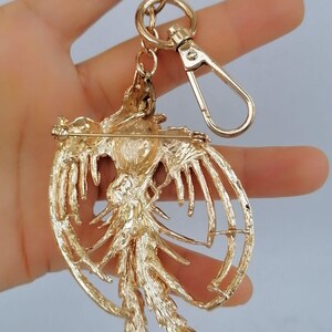 May include: A gold-colored metal keychain featuring a detailed phoenix design. The phoenix has outstretched wings and a long tail, with a pin on the back. The keychain includes a split ring and a lobster clasp for easy attachment.