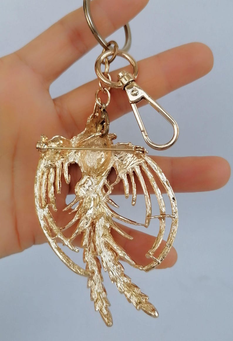 May include: A gold-colored metal keychain featuring a detailed phoenix design. The phoenix has outstretched wings and a long tail, with a pin on the back. The keychain includes a split ring and a lobster clasp for easy attachment.