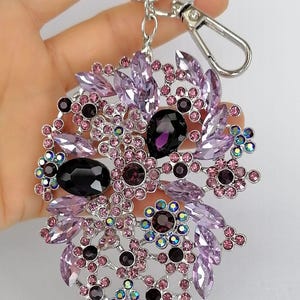 May include: A silver-toned keychain with a large, ornate pendant. The pendant features a floral design with various shades of purple and pink crystals and gemstones. The keychain has a lobster clasp and a key ring.