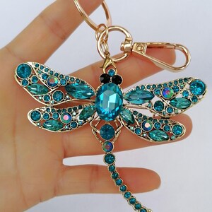 Op de afbeelding: Een goudkleurige sleutelhanger in de vorm van een libel, versierd met teal en blauwe kristallen. De libel heeft een groot centraal kristal en kleinere kristallen die de vleugels en het lichaam omlijnen. De sleutelhanger bevat een sluiting en een ring.