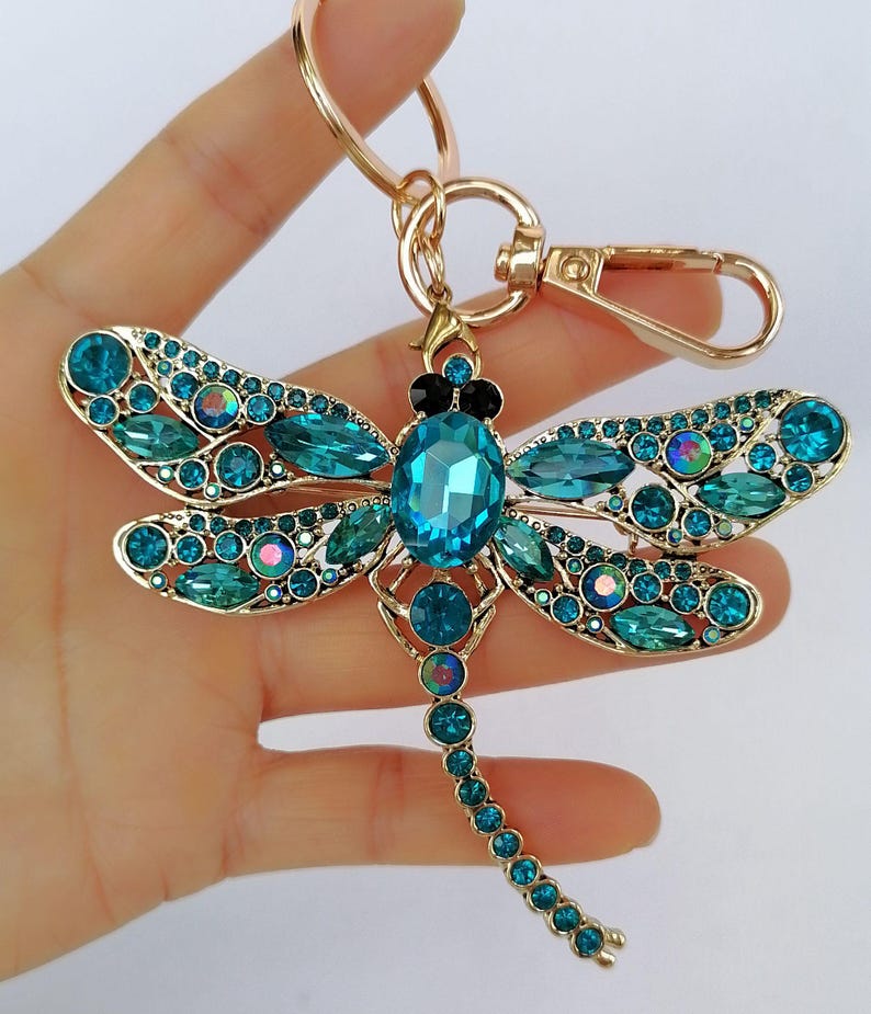 Op de afbeelding: Een goudkleurige sleutelhanger in de vorm van een libel, versierd met teal en blauwe kristallen. De libel heeft een groot centraal kristal en kleinere kristallen die de vleugels en het lichaam omlijnen. De sleutelhanger bevat een sluiting en een ring.