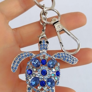 May include: A silver-toned keychain featuring a bejeweled turtle pendant. The turtle is adorned with various shades of blue crystals, creating a sparkling effect. The keychain includes a lobster clasp and a split ring for attaching keys.