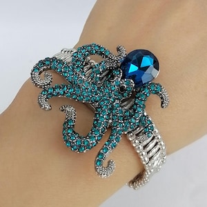 Puede incluir: Una pulsera plateada con un diseño de pulpo turquesa adornado con pedrería turquesa. Una gran gema azul facetada se encuentra sobre la cabeza del pulpo. La pulsera tiene una banda flexible y acanalada.