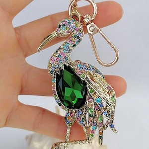 May include: A gold-toned keychain featuring a bejeweled bird design. The bird has a large green gemstone body and is adorned with colorful rhinestones. The keychain includes a clasp and a ring for attaching keys.