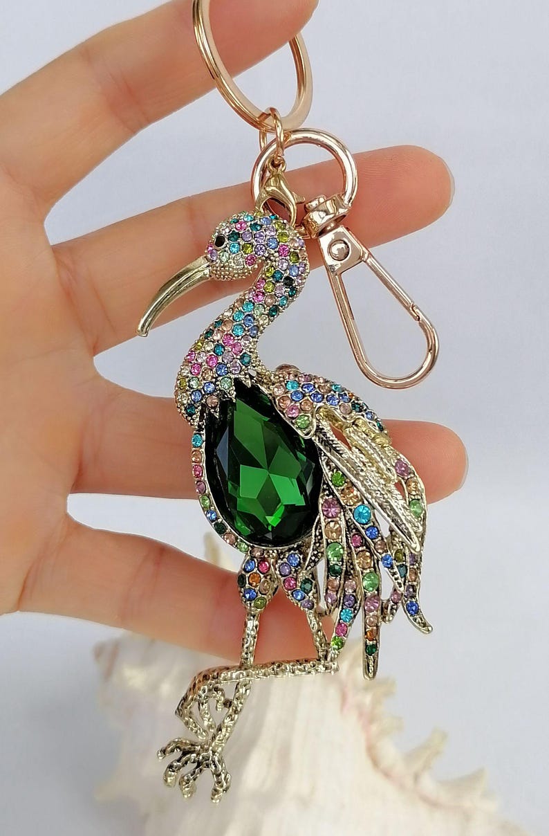 May include: A gold-toned keychain featuring a bejeweled bird design. The bird has a large green gemstone body and is adorned with colorful rhinestones. The keychain includes a clasp and a ring for attaching keys.
