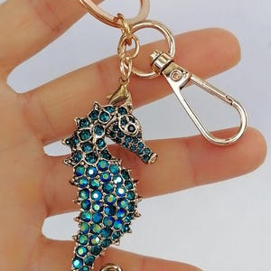 K&ouml;nnte beinhalten: Ein goldfarbener Schl&uuml;sselanh&auml;nger mit einem Seepferdchen-Anh&auml;nger. Das Seepferdchen ist mit blauen und t&uuml;rkisfarbenen Strasssteinen besetzt. Der Schl&uuml;sselanh&auml;nger hat einen Karabinerverschluss und einen Schl&uuml;sselring.