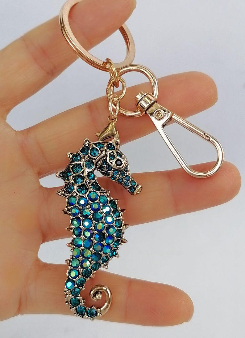 K&ouml;nnte beinhalten: Ein goldfarbener Schl&uuml;sselanh&auml;nger mit einem Seepferdchen-Anh&auml;nger. Das Seepferdchen ist mit blauen und t&uuml;rkisfarbenen Strasssteinen besetzt. Der Schl&uuml;sselanh&auml;nger hat einen Karabinerverschluss und einen Schl&uuml;sselring.