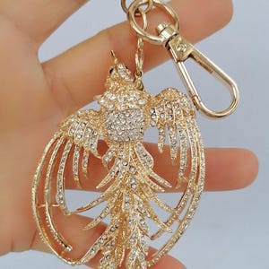 May include: Gold-toned metal keychain featuring a phoenix design, embellished with sparkling rhinestones. The bird's wings are outstretched, and the keychain includes a clasp and ring for attachment. A decorative accessory.