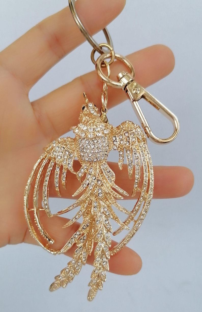 May include: Gold-toned metal keychain featuring a phoenix design, embellished with sparkling rhinestones. The bird's wings are outstretched, and the keychain includes a clasp and ring for attachment. A decorative accessory.