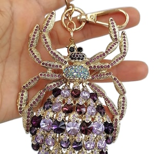May include: Gold-toned spider keychain adorned with purple and iridescent rhinestones. The spider's body features a cluster of purple and lavender gems, with smaller purple gems outlining the legs. A gold clasp and key ring are attached.