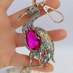 May include: A gold-toned metal keychain featuring a bejeweled bird design. The bird has a large, pink gemstone body and is adorned with colorful rhinestones. The keychain includes a clasp and a ring.