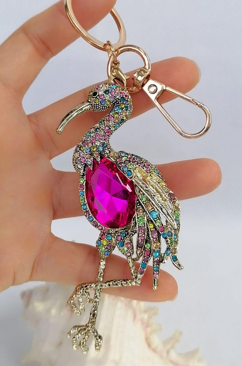May include: A gold-toned metal keychain featuring a bejeweled bird design. The bird has a large, pink gemstone body and is adorned with colorful rhinestones. The keychain includes a clasp and a ring.
