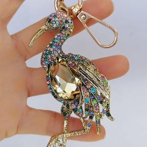May include: A gold-toned bird keychain, adorned with colorful rhinestones. The bird features a large, amber-colored crystal at its center. The keychain includes a clasp and a ring for attaching keys.
