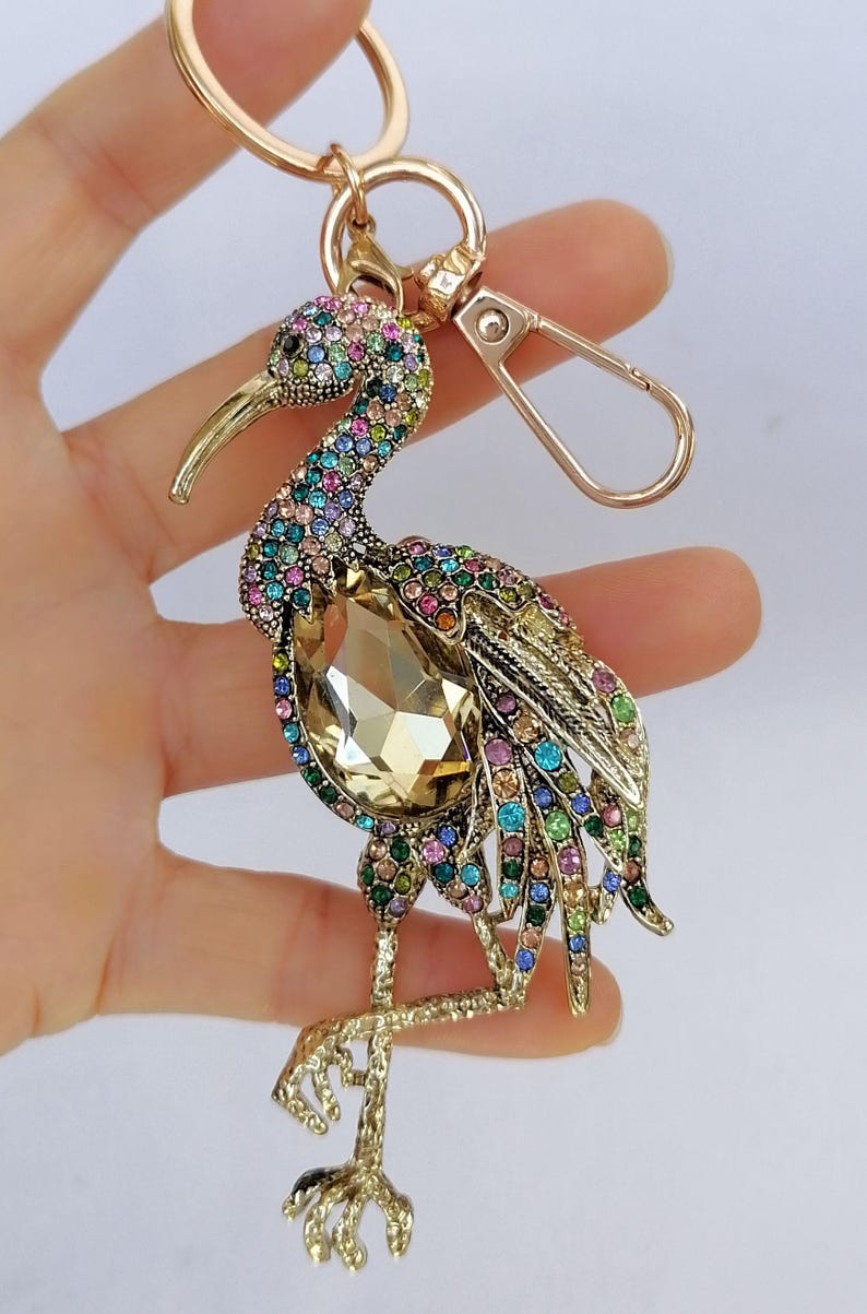 May include: A gold-toned bird keychain, adorned with colorful rhinestones. The bird features a large, amber-colored crystal at its center. The keychain includes a clasp and a ring for attaching keys.