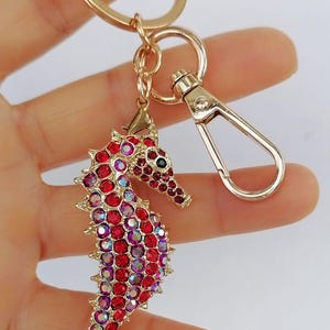 K&ouml;nnte beinhalten: Goldfarbener Seepferdchen-Schl&uuml;sselanh&auml;nger mit roten und irisierenden Strasssteinen. Der Seepferdchen-Anh&auml;nger ist mit einer goldenen Metalloberfl&auml;che und einem Karabinerhaken versehen. Ein dekoratives Accessoire.