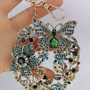 May include: Gold-toned keychain with a butterfly and floral design. The butterfly has teal-colored jewel accents, and a large green jewel at the center. The floral elements are also adorned with teal and green jewels.