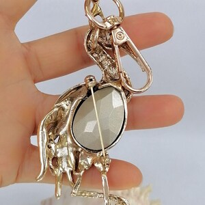 May include: Gold-toned metal keychain featuring a stylized bird design. The bird has a large, faceted, oval-shaped, gray stone set in its body. The keychain includes a clasp and a key ring.