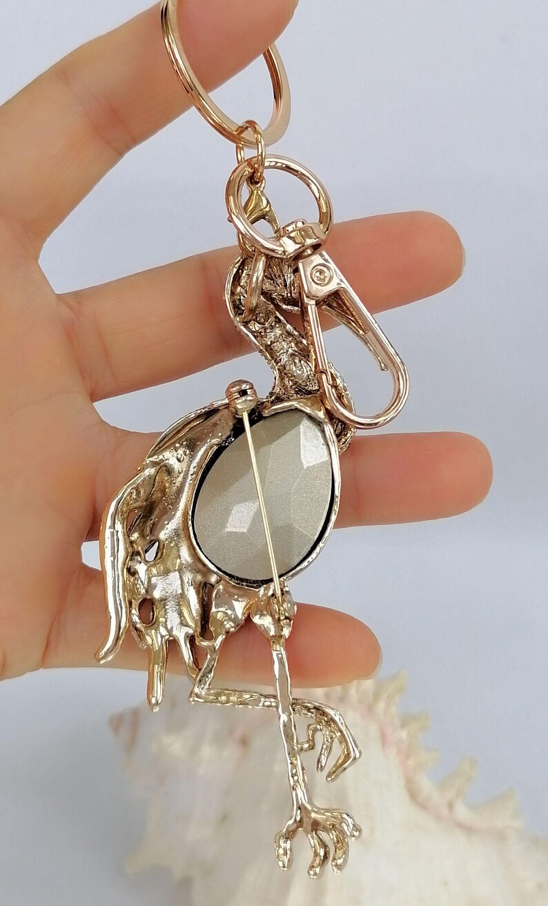 May include: Gold-toned metal keychain featuring a stylized bird design. The bird has a large, faceted, oval-shaped, gray stone set in its body. The keychain includes a clasp and a key ring.