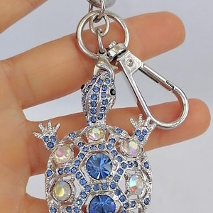 Puede incluir: Llavero de tortuga de metal plateado con detalles de cristales azules e iridiscentes. El caparazón de la tortuga presenta un diseño geométrico con grandes cristales azules. Incluye un llavero y un mosquetón.