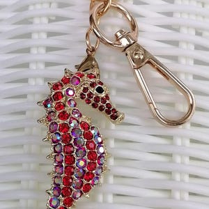 K&ouml;nnte beinhalten: Ein goldfarbener Schl&uuml;sselanh&auml;nger in Form eines Seepferdchens mit roten und irisierenden Strasssteinen. Das Seepferdchen hat ein detailliertes Design und ist an einem goldfarbenen Schl&uuml;sselring und Verschluss befestigt. Der Hintergrund ist ein wei&szlig;es Webmuster.
