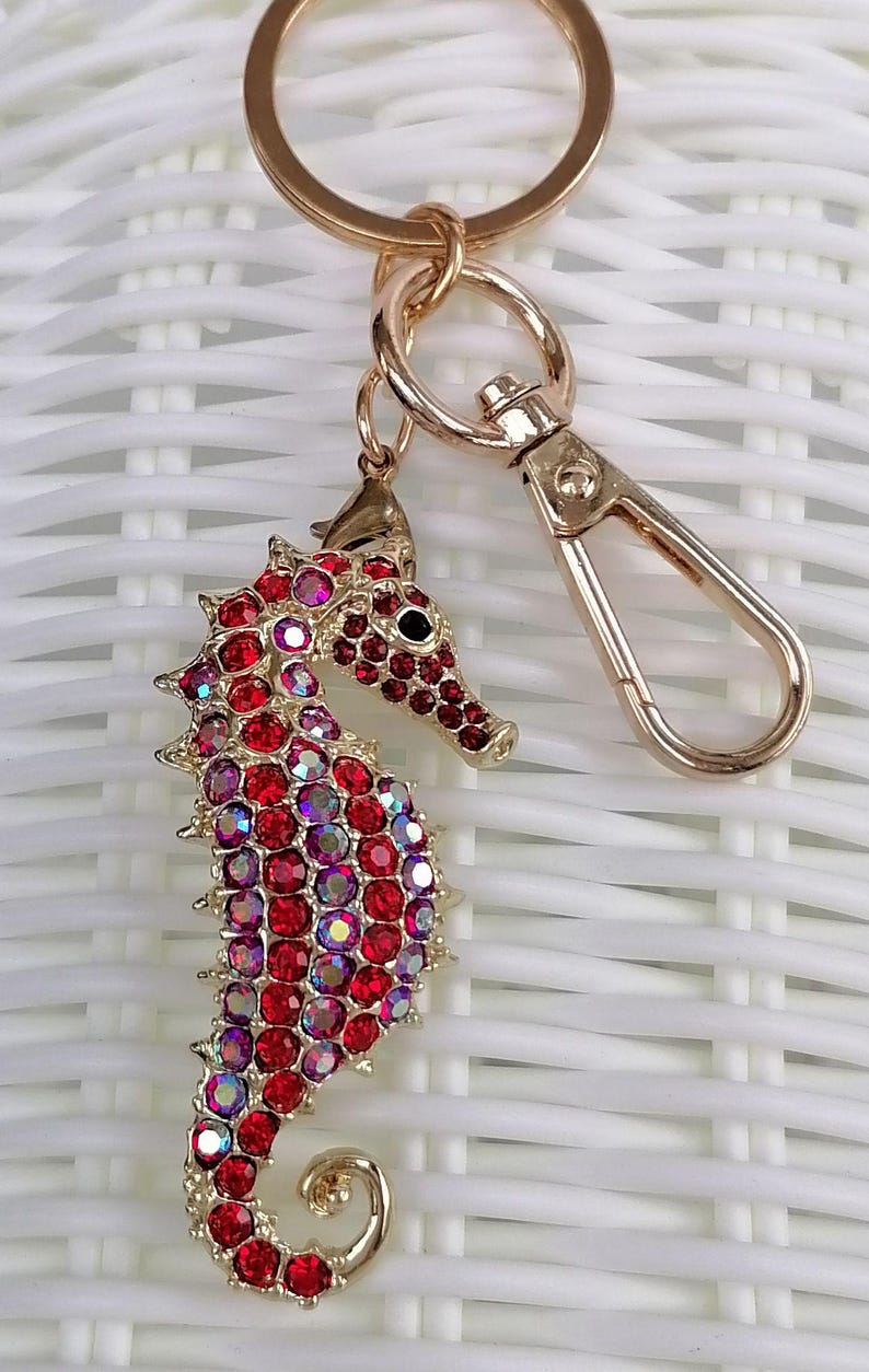 K&ouml;nnte beinhalten: Ein goldfarbener Schl&uuml;sselanh&auml;nger in Form eines Seepferdchens mit roten und irisierenden Strasssteinen. Das Seepferdchen hat ein detailliertes Design und ist an einem goldfarbenen Schl&uuml;sselring und Verschluss befestigt. Der Hintergrund ist ein wei&szlig;es Webmuster.