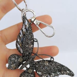 May include: A silver-toned butterfly keychain with a lobster clasp and key ring. The butterfly is encrusted with gray rhinestones, creating a sparkling effect. The wings and body are detailed with various sizes of stones.
