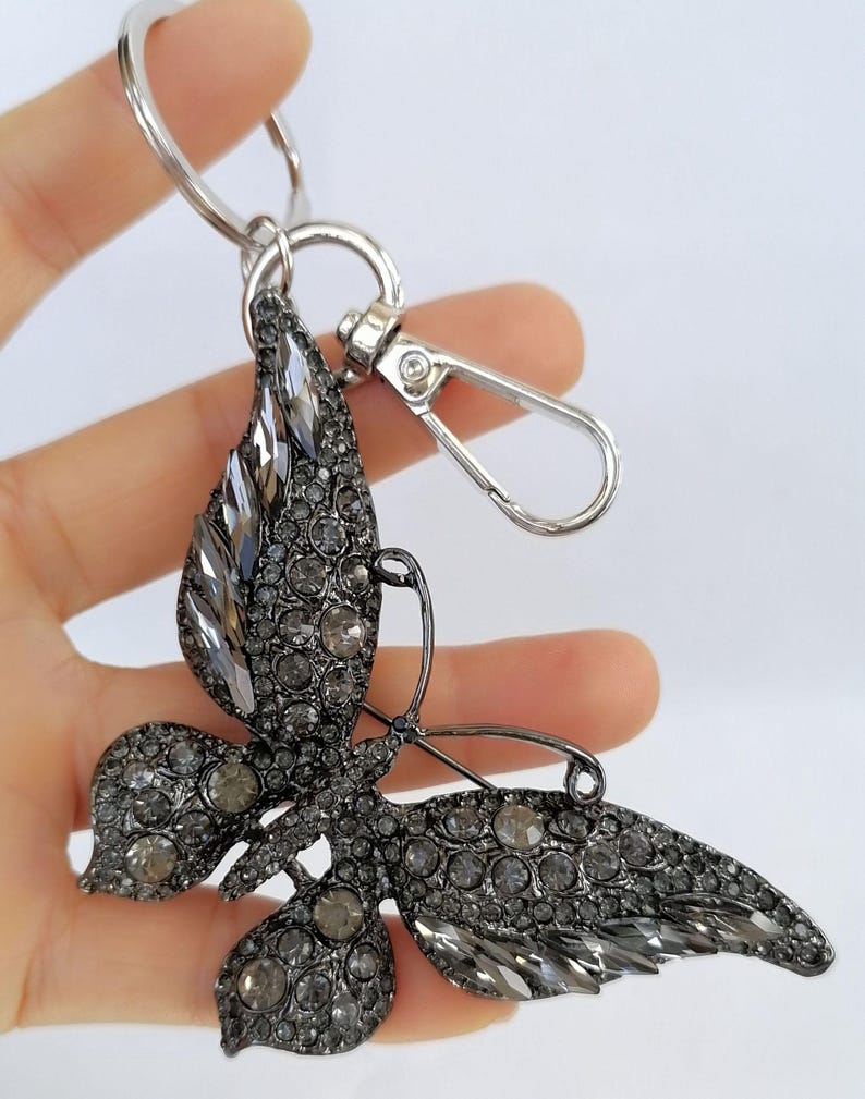 May include: A silver-toned butterfly keychain with a lobster clasp and key ring. The butterfly is encrusted with gray rhinestones, creating a sparkling effect. The wings and body are detailed with various sizes of stones.