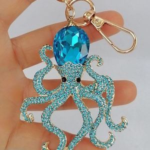 May include: A gold-toned metal keychain featuring a bejeweled octopus design. The octopus has turquoise-colored rhinestones and a large, faceted blue crystal head. The keychain includes a lobster clasp and a key ring.