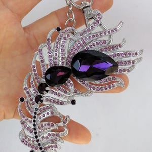 May include: Silver-toned keychain with a decorative feather and jewel design. The feather is encrusted with small pink crystals, and the focal point features two large, dark purple teardrop-shaped jewels. A stylish accessory.