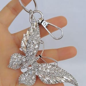 May include: A silver-toned butterfly keychain, encrusted with clear crystals. The butterfly has detailed wings and a metal clasp for attaching to keys or a bag. The keychain is held in a hand against a white background.