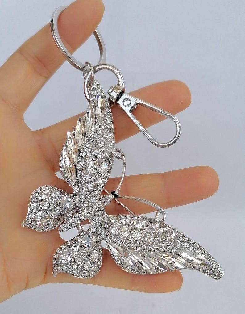 May include: A silver-toned butterfly keychain, encrusted with clear crystals. The butterfly has detailed wings and a metal clasp for attaching to keys or a bag. The keychain is held in a hand against a white background.