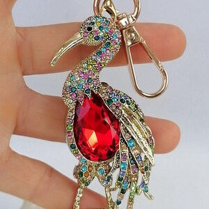 May include: A gold-toned metal keychain featuring a bejeweled bird design. The bird has a large red teardrop-shaped gem on its chest and is adorned with colorful rhinestones. The keychain includes a clasp and a ring.