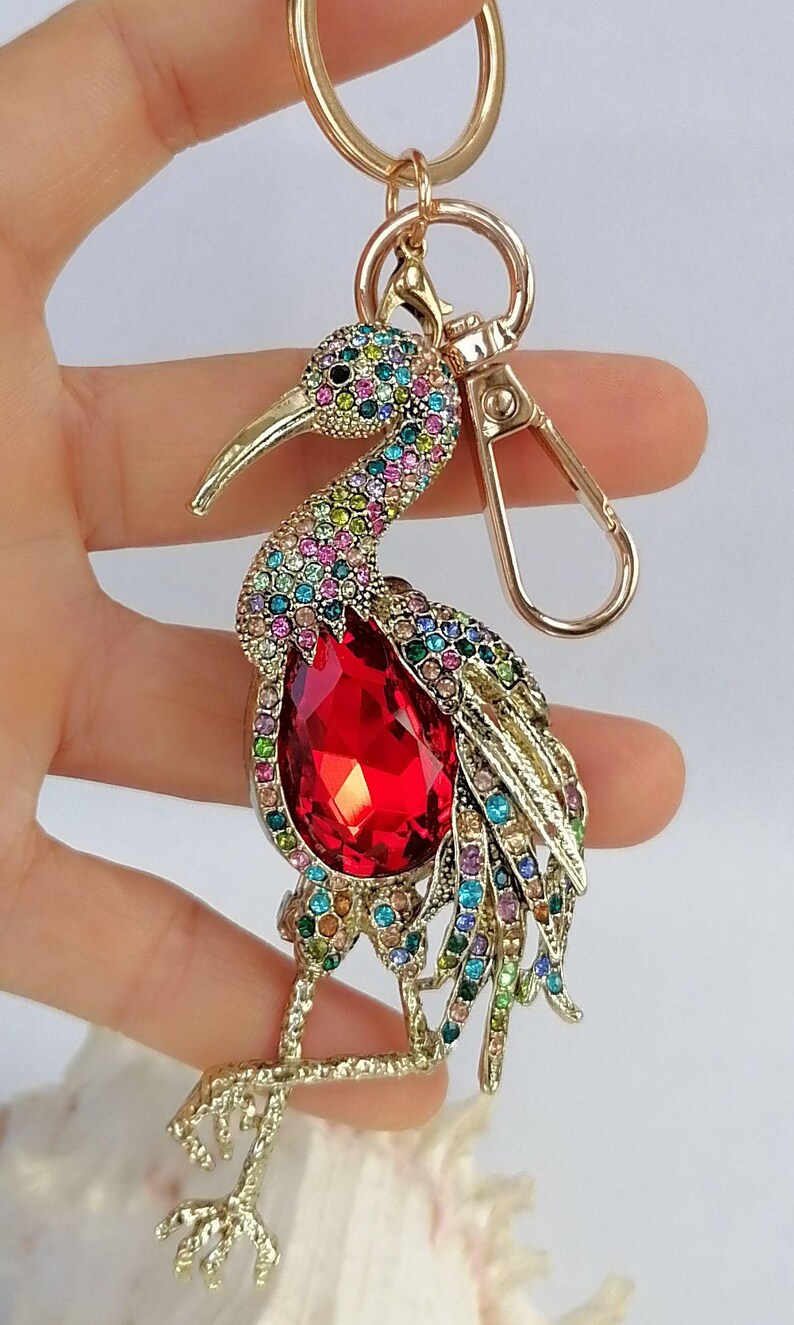 May include: A gold-toned metal keychain featuring a bejeweled bird design. The bird has a large red teardrop-shaped gem on its chest and is adorned with colorful rhinestones. The keychain includes a clasp and a ring.