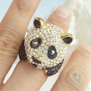 May include: A gold-toned ring shaped like a panda's head, encrusted with clear rhinestones. The panda has black ears, eyes, and nose, with a black and rhinestone-covered lower jaw. The ring is a fun and whimsical accessory.