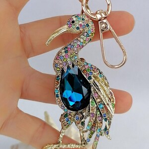 May include: A bejeweled bird keychain with a gold-tone finish. The bird features a large, teardrop-shaped, dark blue crystal and is adorned with colorful rhinestones. The keychain includes a gold ring and clasp.