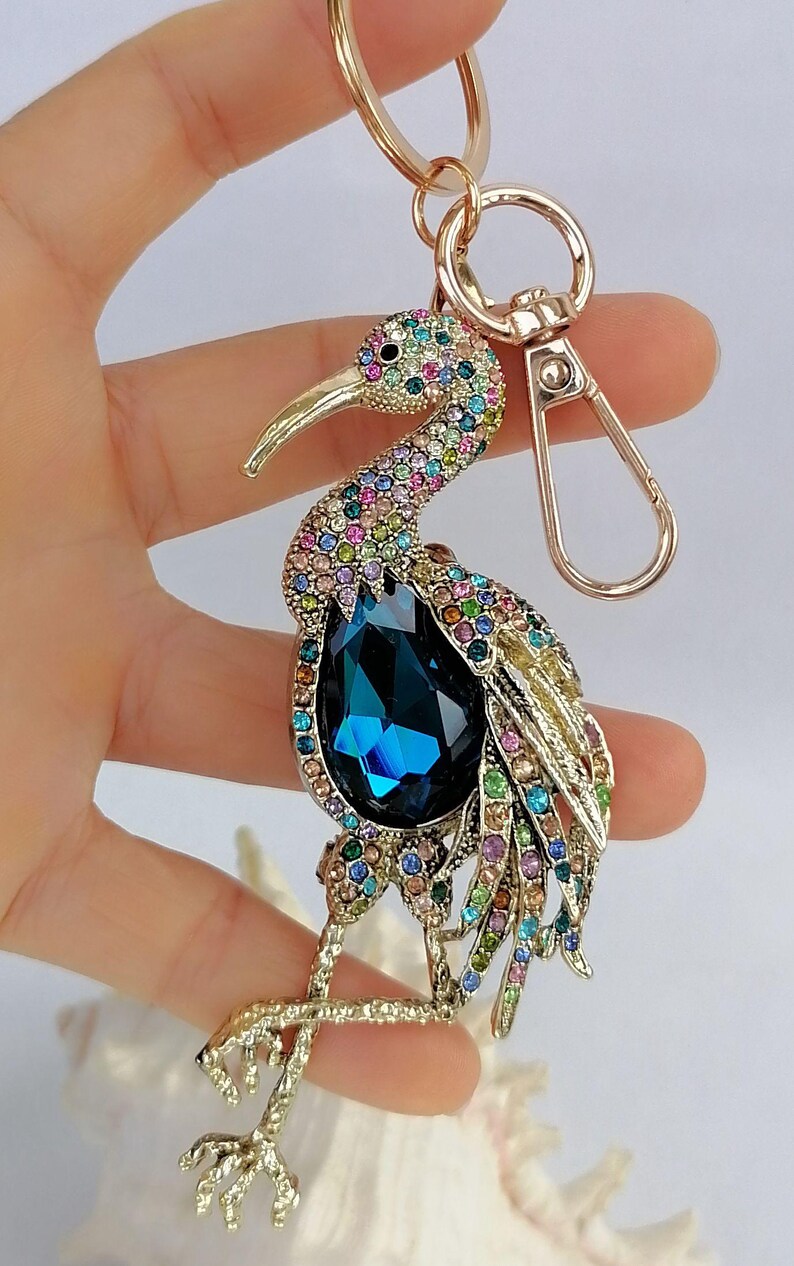 May include: A bejeweled bird keychain with a gold-tone finish. The bird features a large, teardrop-shaped, dark blue crystal and is adorned with colorful rhinestones. The keychain includes a gold ring and clasp.