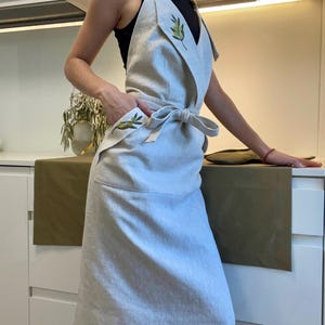 Puede incluir: Delantal de lino beige claro con bordado de ramas de olivo. El delantal presenta un diseño envolvente con cierre de lazo en la cintura y un bolsillo en la parte delantera. El diseño de la rama de olivo está bordado en hilo verde.