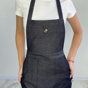 May include: Dark denim apron with a large front pocket and a decorative button closure. The apron has a bib top with long straps and two lower pockets. The apron is designed to be worn over clothing.