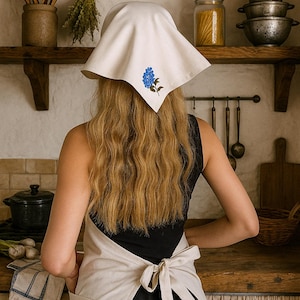 Könnte beinhalten: Eine Frau in einer Küche trägt ein weißes Kopftuch mit blauer Blumenstickerei und eine beige Schürze, die an der Taille gebunden ist. Die Küche hat Holzregale mit Kochutensilien und Gläsern mit Lebensmitteln. Die Frau steht dem Tresen zugewandt.