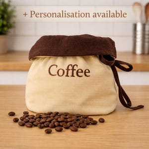 May include: A cream-coloured fabric storage bag with a dark brown top cuff and drawstring closure. The word Coffee is embroidered in dark brown on the front. A pile of roasted coffee beans is scattered in front of the bag. Text reads + Personalisation available.