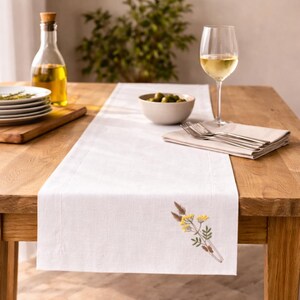 May include: A white linen table runner featuring an embroidered botanical design of yellow flowers and brown grasses. The runner is laid flat on a wooden table, accompanied by a glass of white wine, a bowl of olives, and a stack of white plates.
