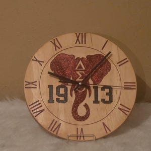 May include: A round wooden clock with Roman numerals and black hands. The clock face features a red glitter elephant design with the numbers "1913" and a Greek symbol. The clock is displayed on a white surface.
