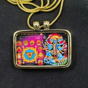 Puede incluir: Un collar con colgante rectangular de tono dorado que presenta una figura de deidad azul de múltiples brazos a la derecha y un diagrama yantra geométrico colorido a la izquierda, rodeado de iconos de deidades más pequeños en un marco de metal dorado.