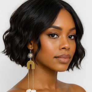May include: A woman with dark hair and skin wears long, dangling earrings. The earrings feature a sunburst, a gold arch, and a cluster of white, organic-shaped beads. The earrings hang from a small hoop.