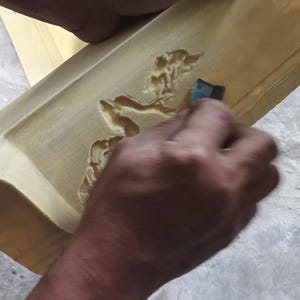 May include: A person is sanding a light brown wooden object with a carved design. The object appears to be a tray or a similar item. The image is taken from a close-up angle, showing the details of the wood and the sanding process.